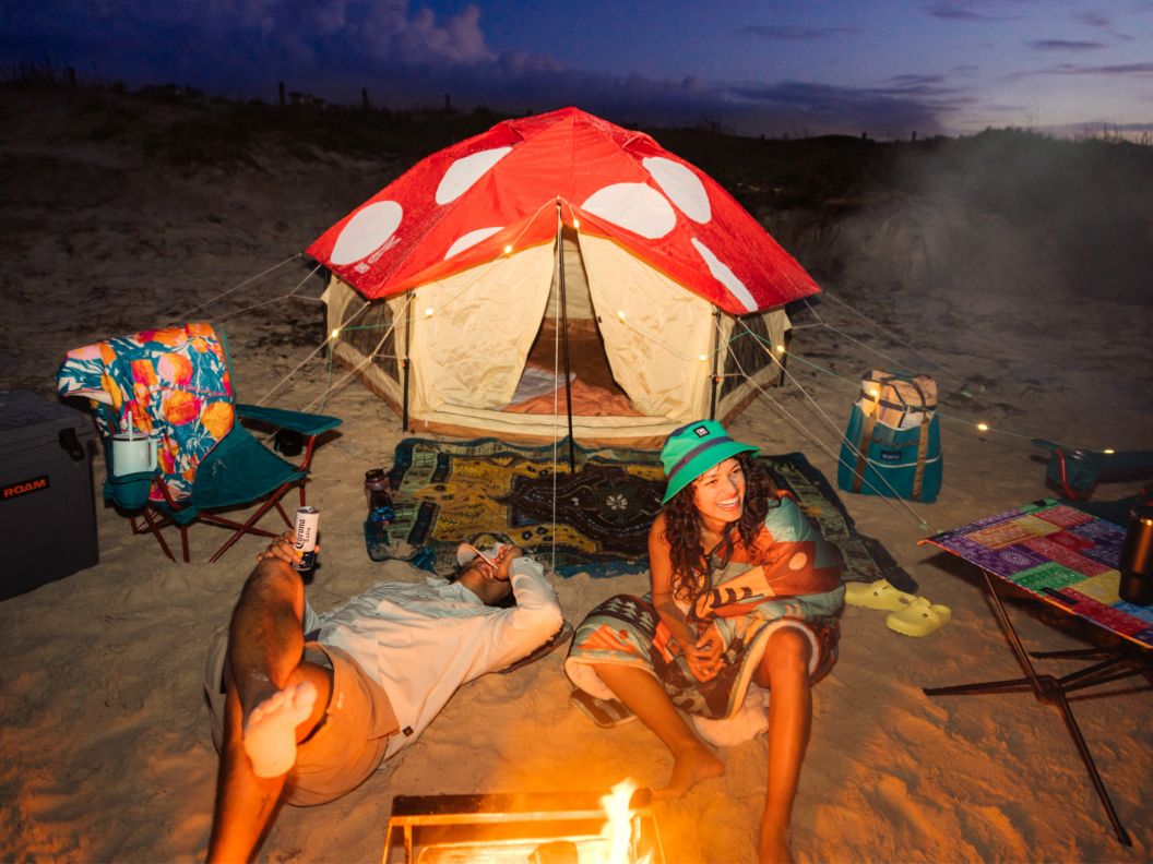 Two people partying at a campsite with chairs, a tent, and fire pit set up on a beach.